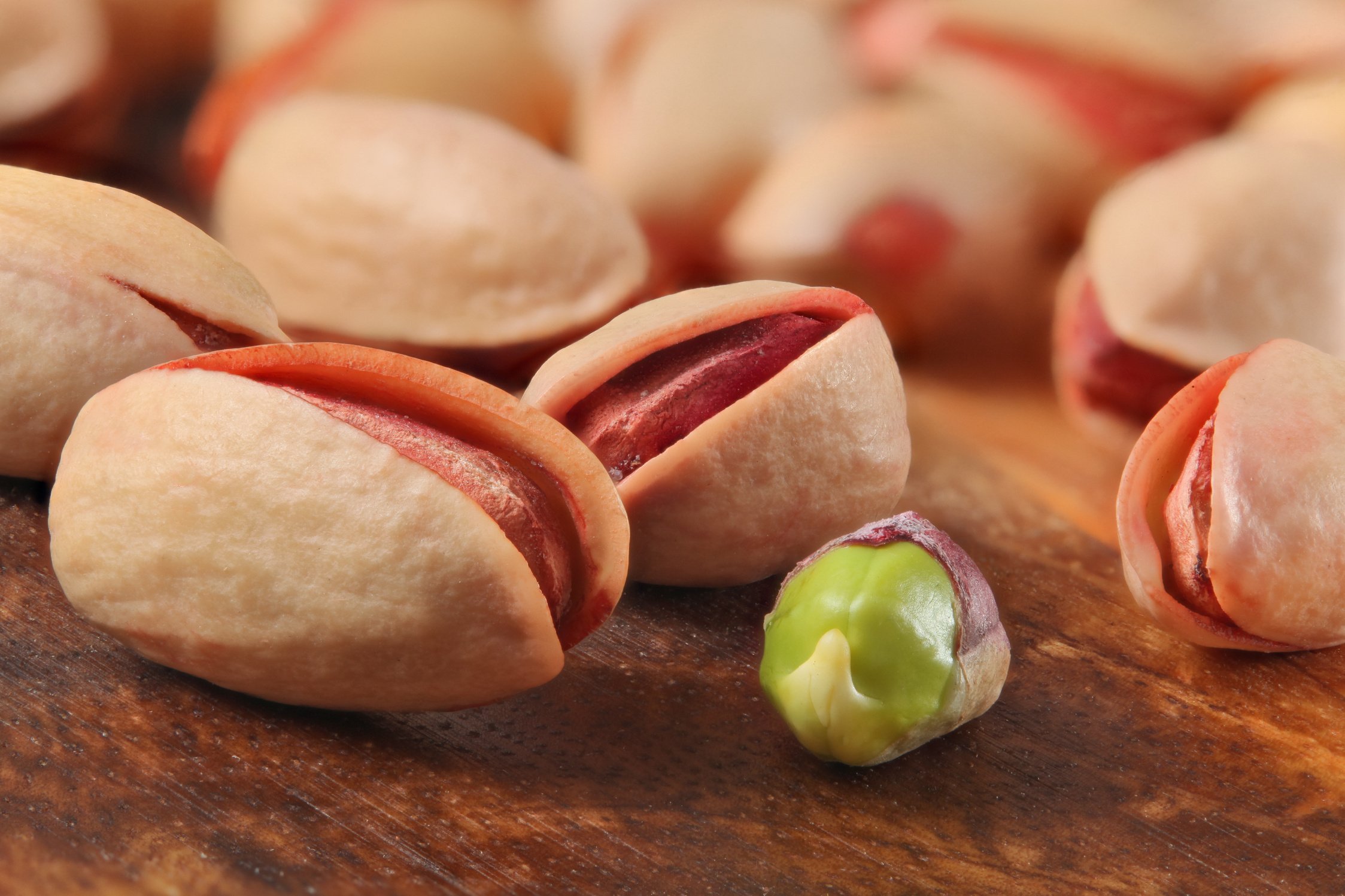 Roasted salted Turkish red pistachios, one of them peeled, green seed visible, on wooden board. Closeup detail.